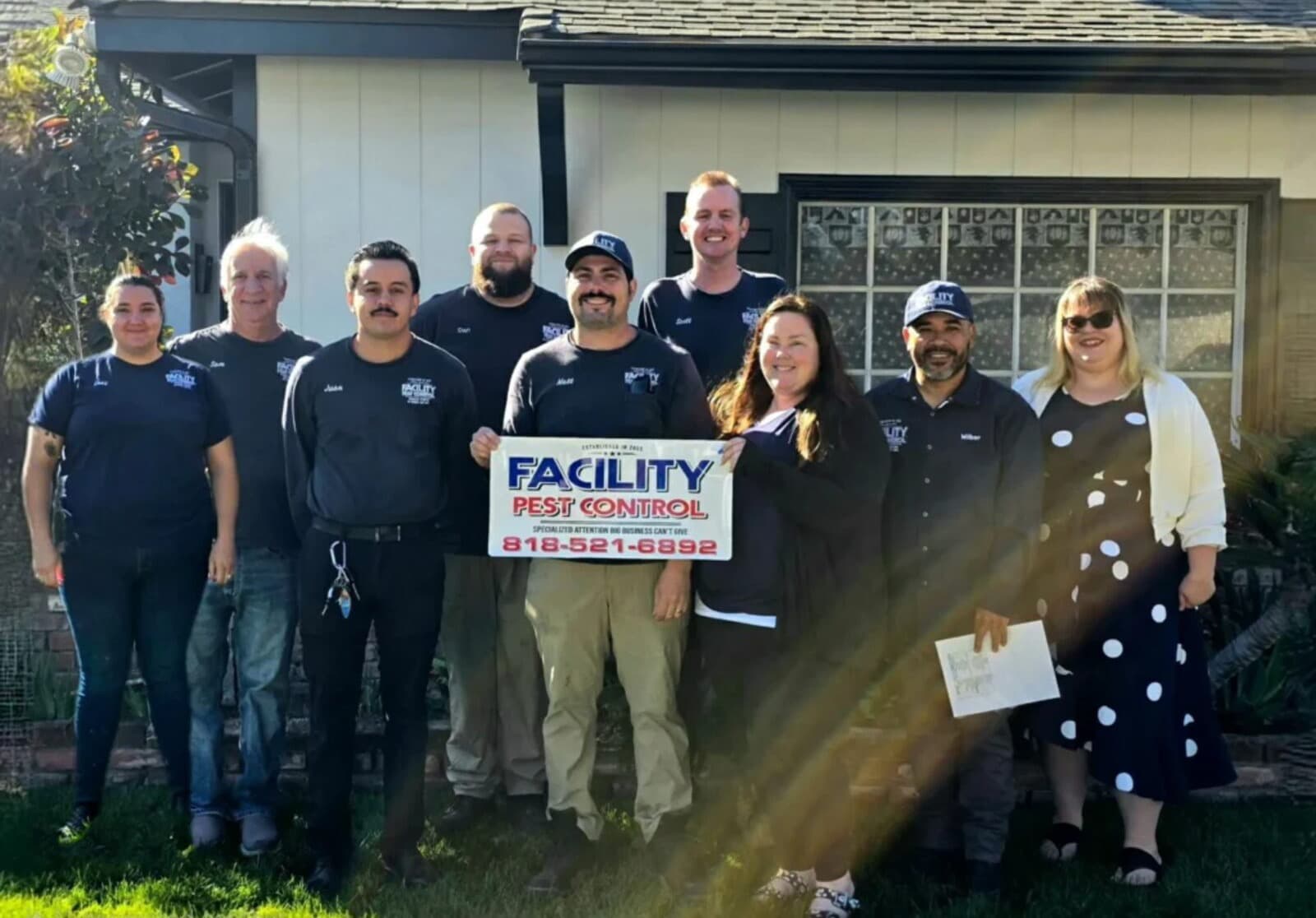The Facility Pest Control team of nine in navy uniform shirts in front of a Reseda home, holding the company sign